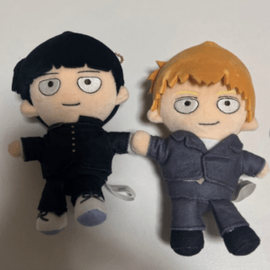 Two plush dolls lying on their backs, holding hands; one has black hair and a black outfit, the other has blond hair and a blue-gray suit, both with light skin and simple facial features on a white background.