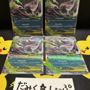 Four holographic Pokémon cards arranged in a 2x2 grid on a dark surface, each featuring a dragon-like figure; a handwritten Japanese sign sits in front.