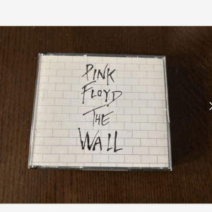CD case featuring a white brick-wall design with handwritten 'Pink Floyd The Wall' on a dark wooden table surface