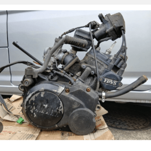 Detached motorcycle engine lying on cardboard outside, with hoses, wiring, and spark plug visible.
