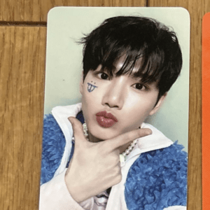 Photocard of a young man with dark hair, wearing a blue textured jacket over white, making a puckered-lip pose with a blue doodle on his left cheek.