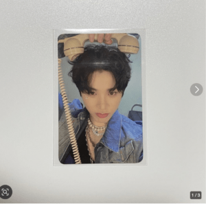 Portrait of a young person with short dark hair wearing a denim jacket and layered necklaces, posing with a retro beige telephone held above their head against a light blue background