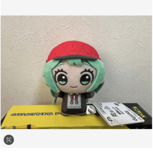 Plush toy of a cute anime-style girl with teal hair and a red cap, standing on a yellow box against a textured wall.