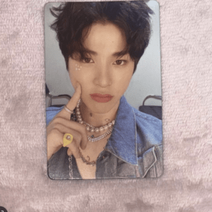 Person with short dark hair wearing a denim jacket and layered necklaces, touching their cheek with a finger while looking at the camera, posed on a pink surface.