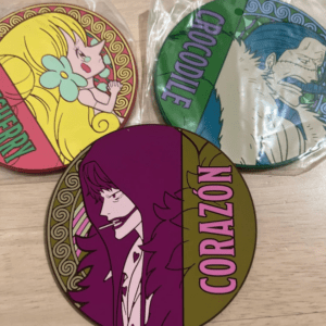 Three round collectible pins on a wooden surface, featuring anime characters: a purple-haired man labeled 'CORAZON' in the foreground, a yellow-haired girl with a flower on the left, and a blue-haired character with 'CROCODILE' on the green pin in the back (some pins still in plastic).  Articulos Japan (Post Box Japan)
